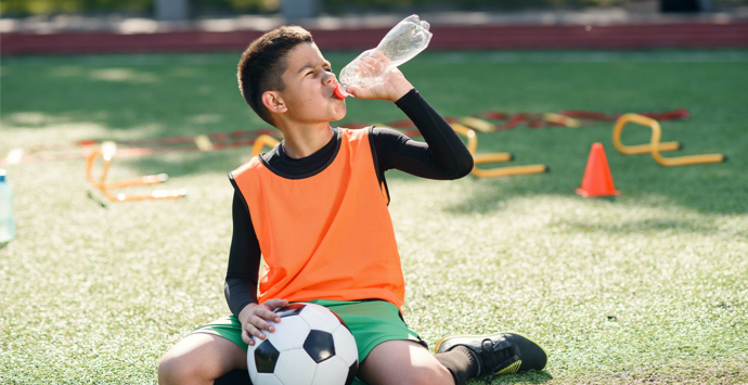 Boy in soccer uniform drinks water from plastic bottle after intensive training at stadium