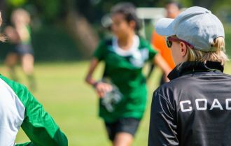 female soccer, football, coach in black coach shirt standing in the sun watching her team play at an outdoor football field